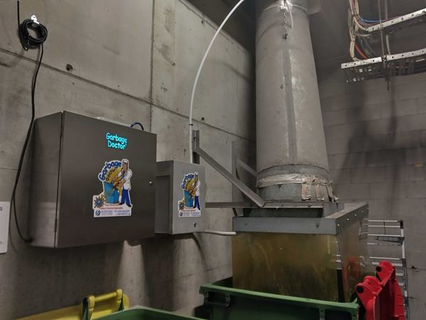 A Garbage Doctor Duo on a bin room wall beside the chute's discharge, its scent tank between the main unit and the chute, above a green bin