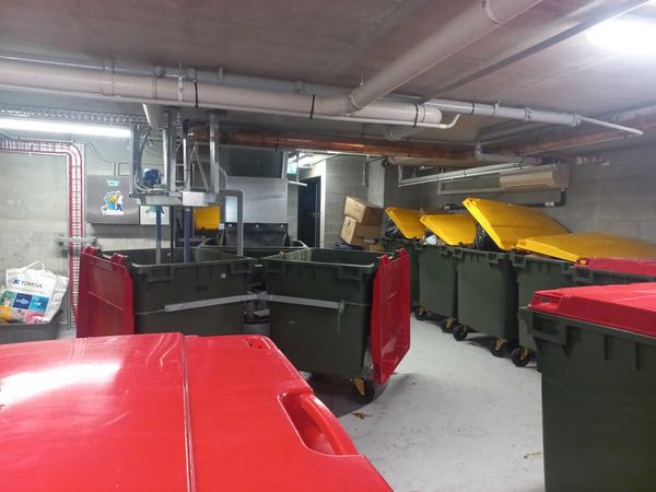 A large basement bin room with its chute, rows of green bins with red and yellow lids, and a Garbage Doctor unit on the far wall