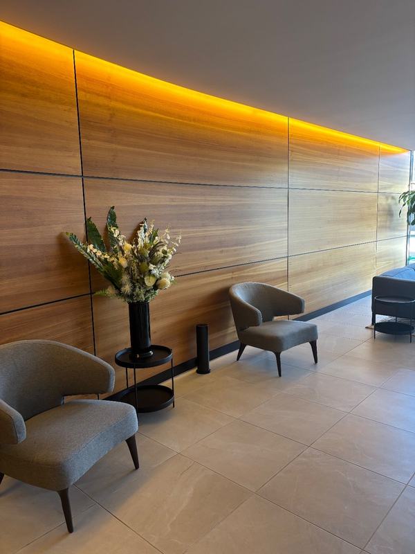 A slim black Scent Doctor tower on the floor of a lobby, beside a vase of flowers and two armchairs, against a timber-panelled wall